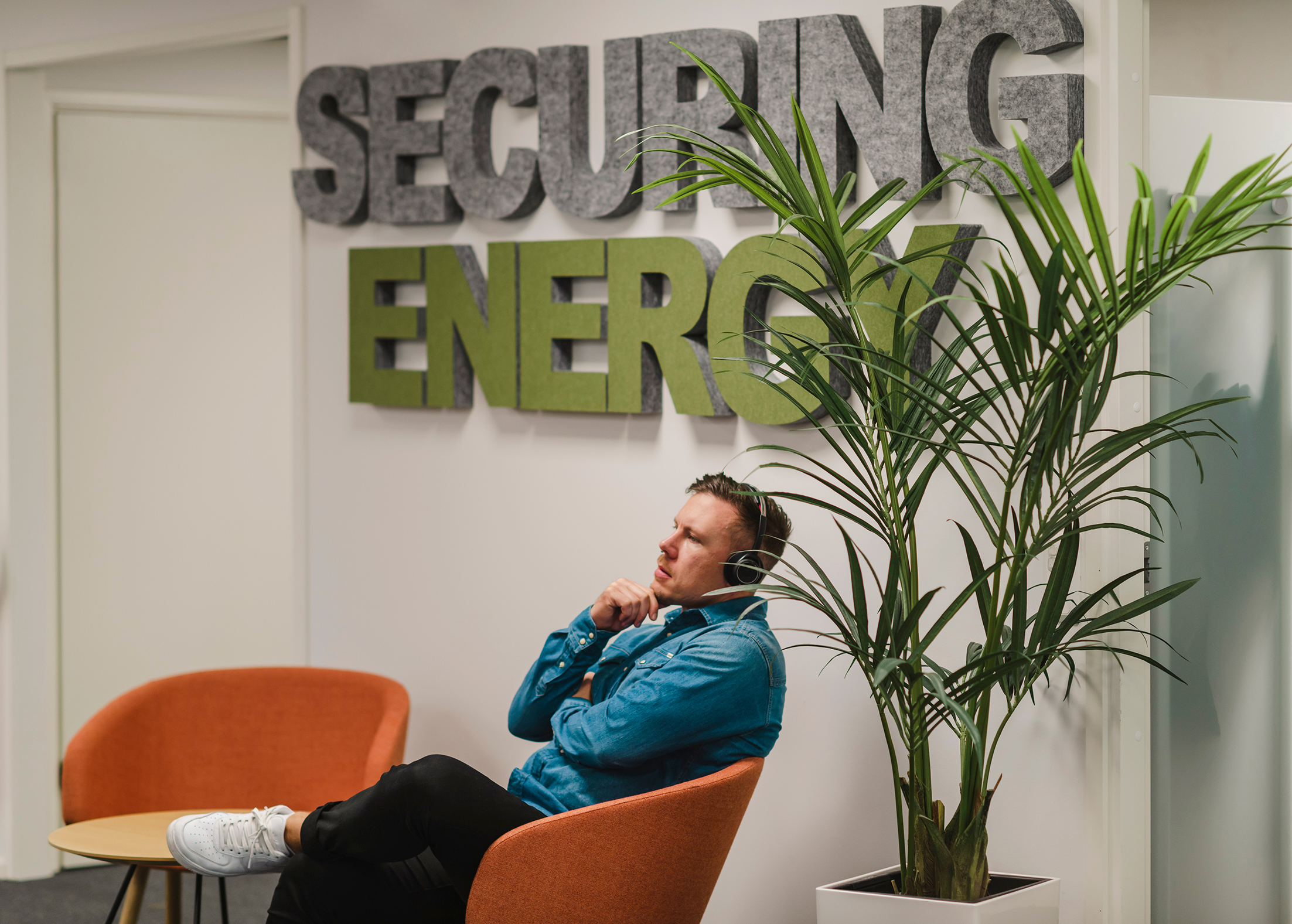 En person sitter i en orange stol i ett kontorsutrymme. En skylt bakom visar texten "SECURING ENERGY," och en hög krukväxt står i närheten.