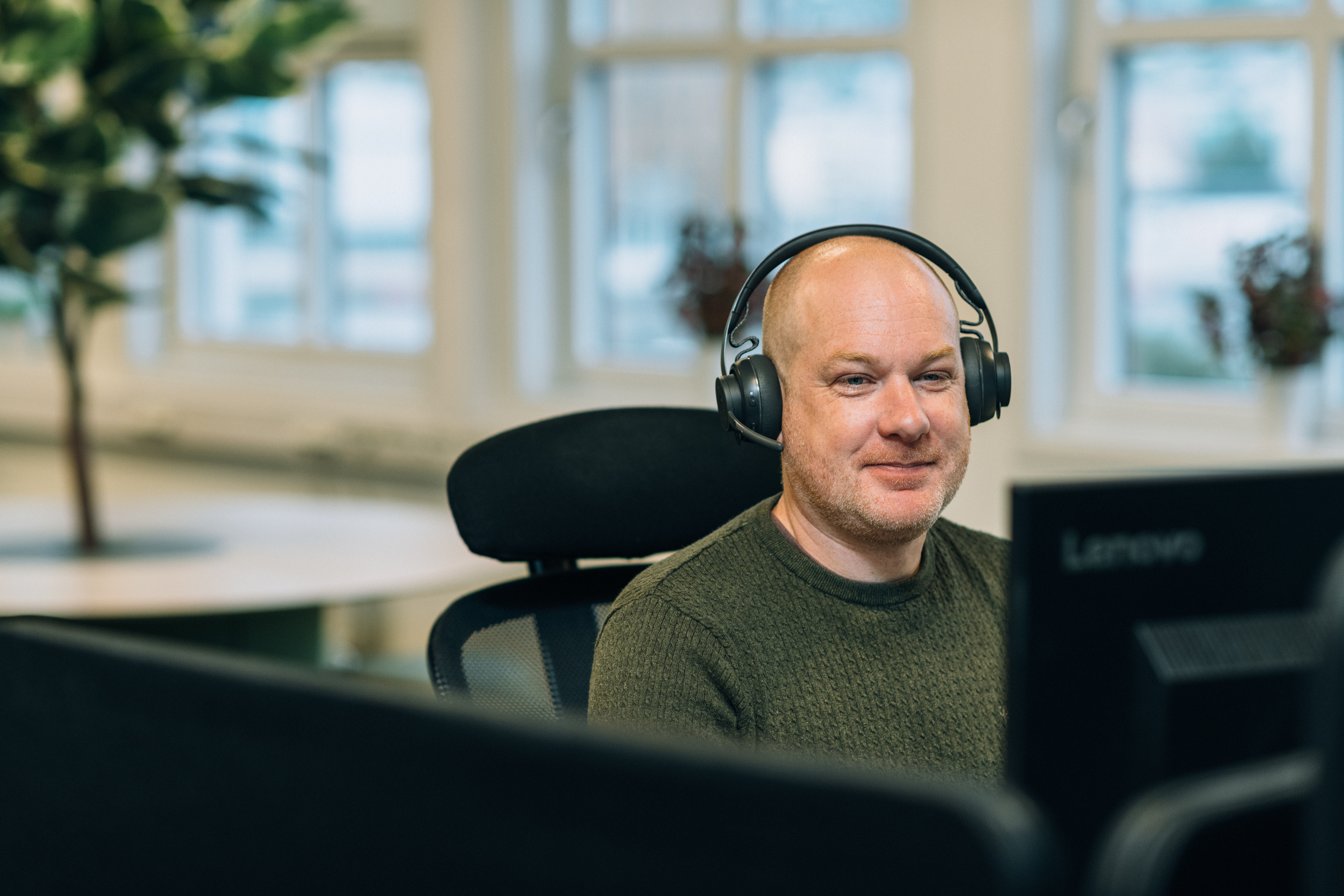 Person med hörlurar tittar på en Lenovo-skärm på ett kontor, med fönster och växter i bakgrunden.