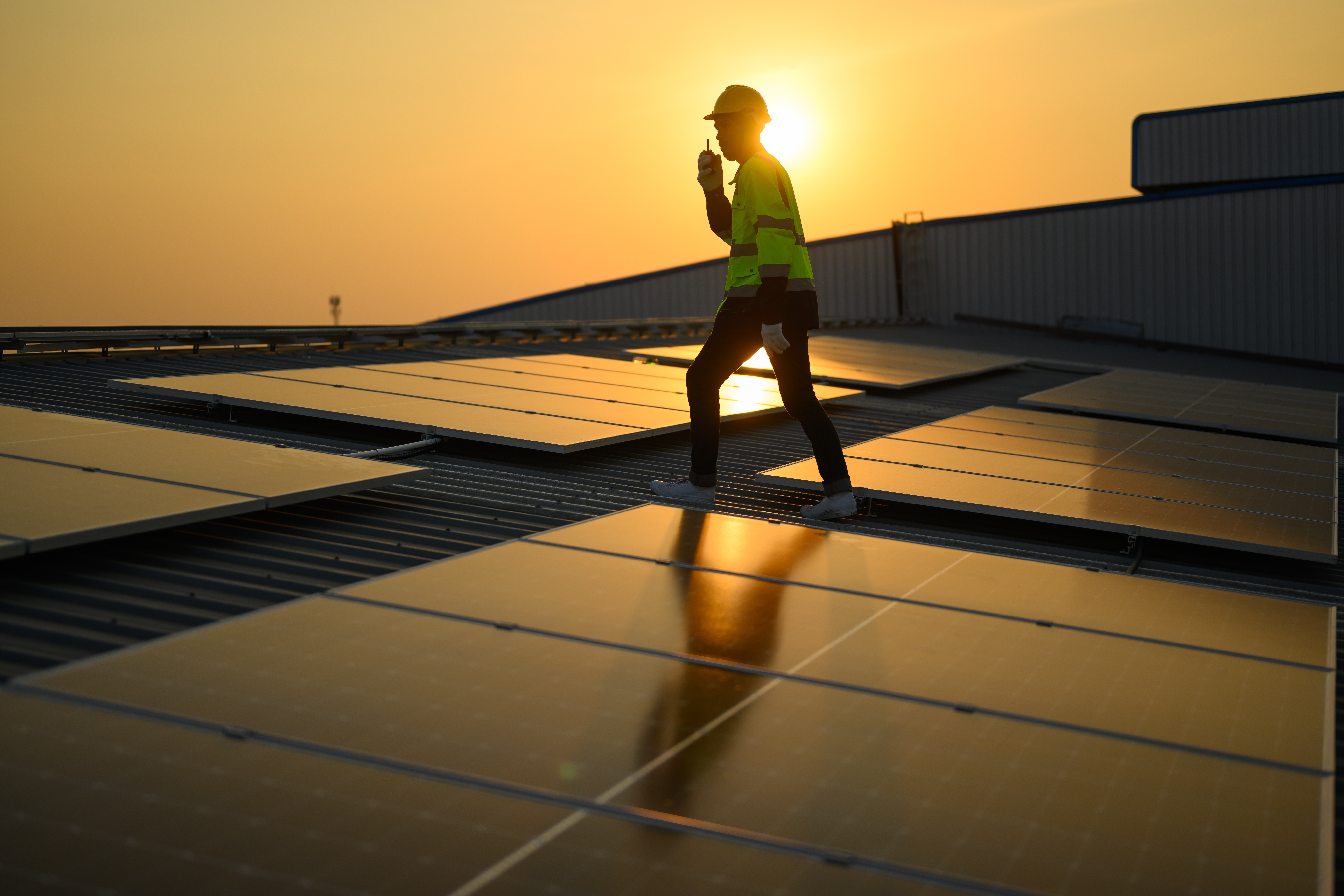 Person med skyddsutrustning går på ett tak med solpaneler vid solnedgången.