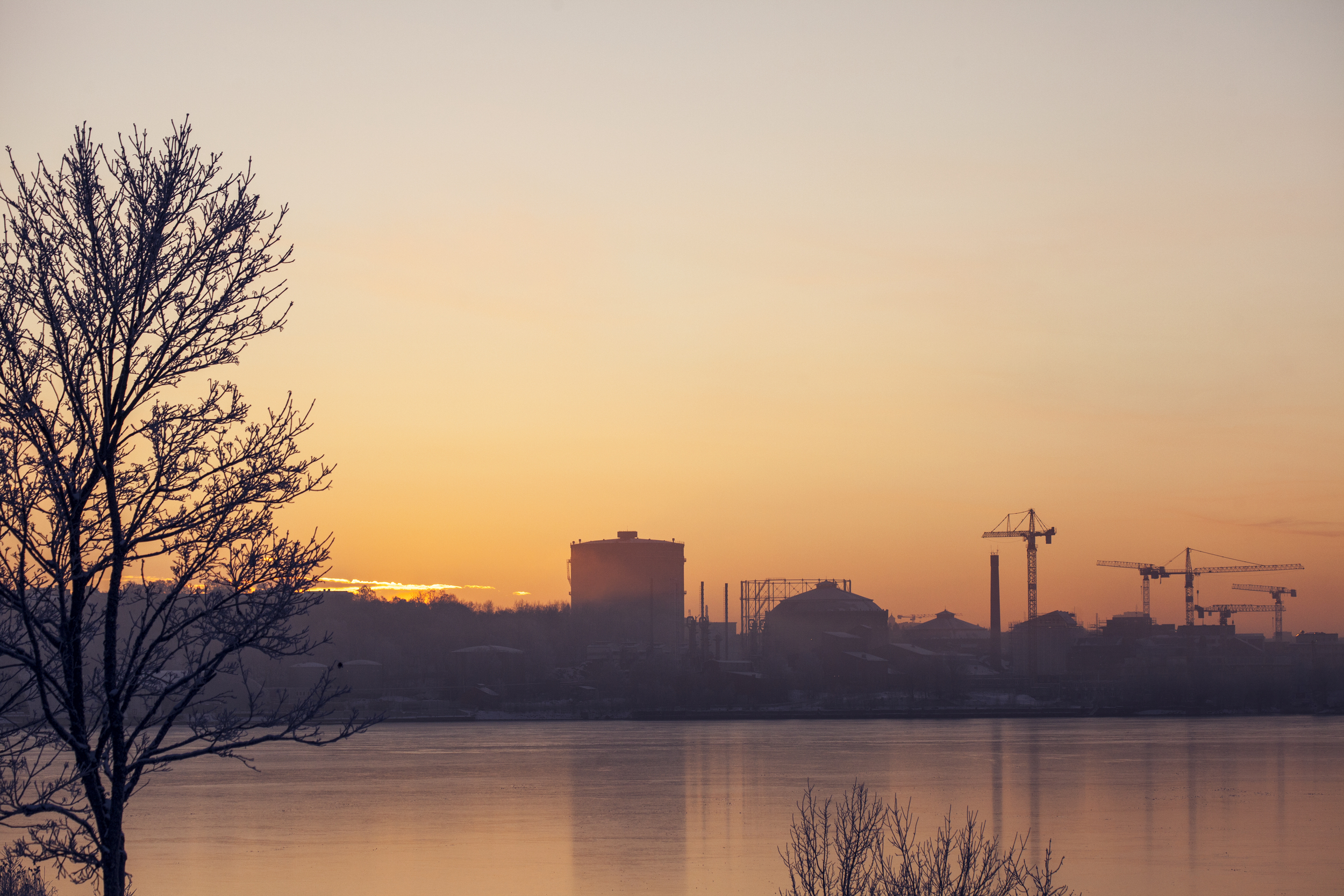 Solnedgång eller soluppgång över vatten med silhuett av träd och en industriell horisont i bakgrunden.